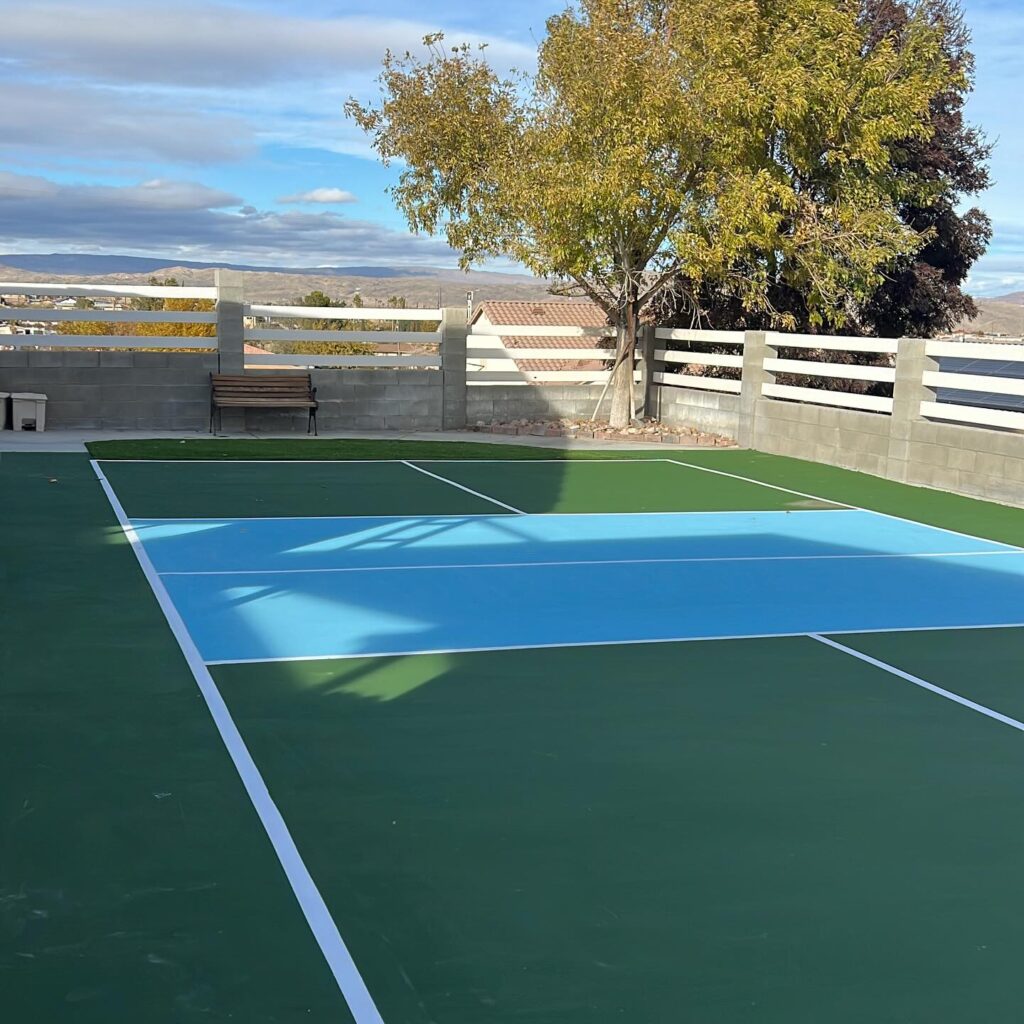 A newly installed backyard pickleball court with a green and blue surface, surrounded by a concrete block wall with a white fence, a wooden bench, and a large tree, under a blue sky.