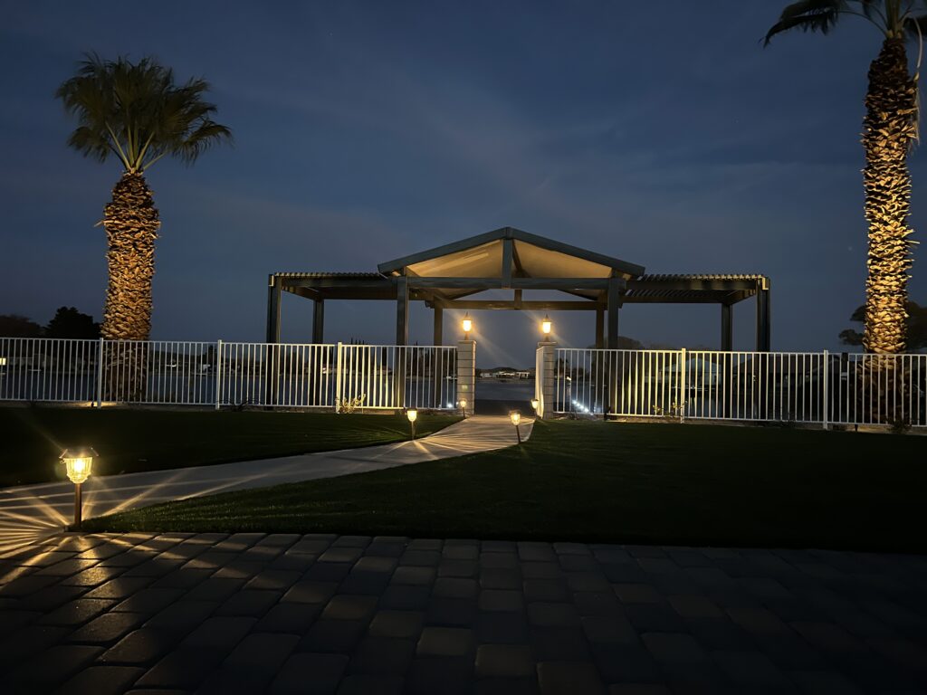 A nighttime view of a landscaped backyard in Silver Lakes, CA, featuring a lit gazebo by the water, palm trees with uplighting, a paved walkway, and manicured turf.
