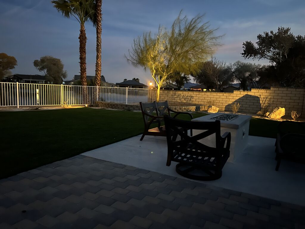 A backyard at twilight featuring artificial turf, a paver patio with a fire pit, and accent landscape lighting against a stone wall.