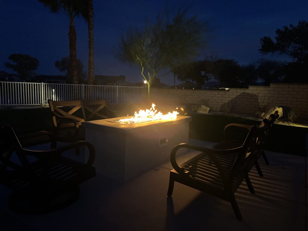 An outdoor fire pit feature glowing at night on a backyard patio, surrounded by chairs with palm trees and a white fence in the background.