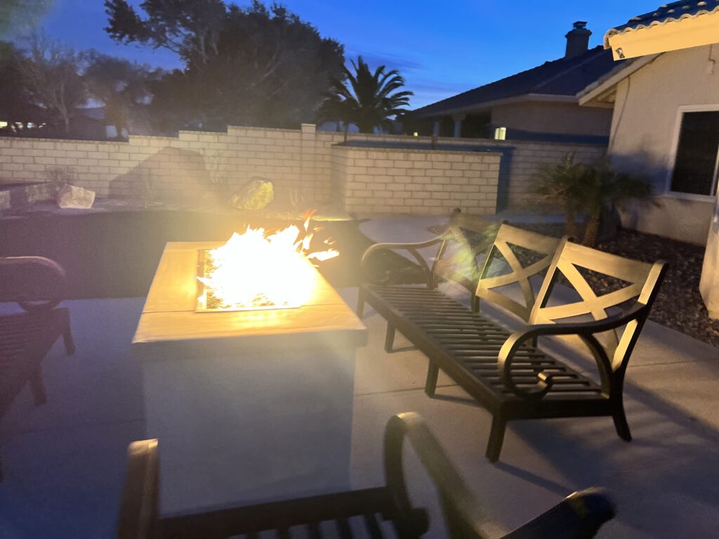 A backyard patio at twilight featuring a glowing rectangular gas fire pit and outdoor seating benches.