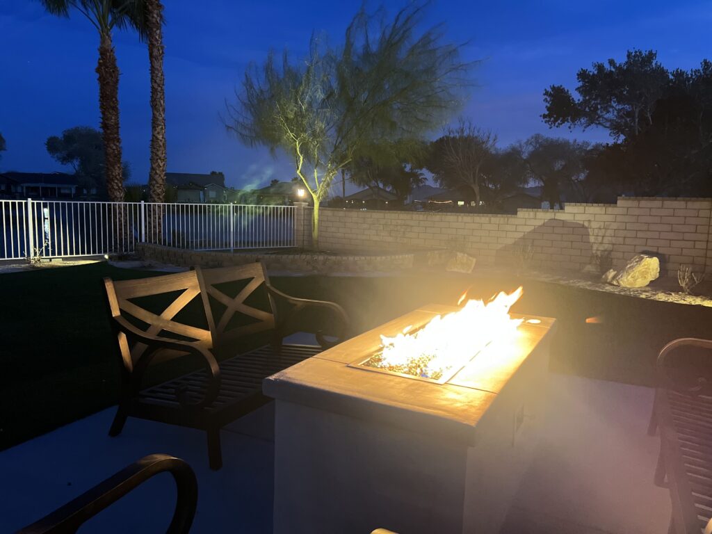 A backyard patio at twilight featuring a rectangular fire pit with flames, comfortable wooden seating, and soft landscape lighting against a dark blue evening sky.