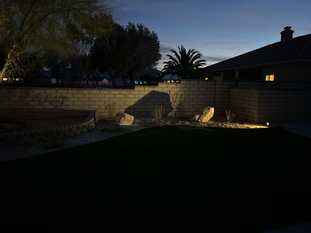 A nighttime view of a backyard featuring landscape lighting accenting stone boulders and a retaining wall against a block wall fence.