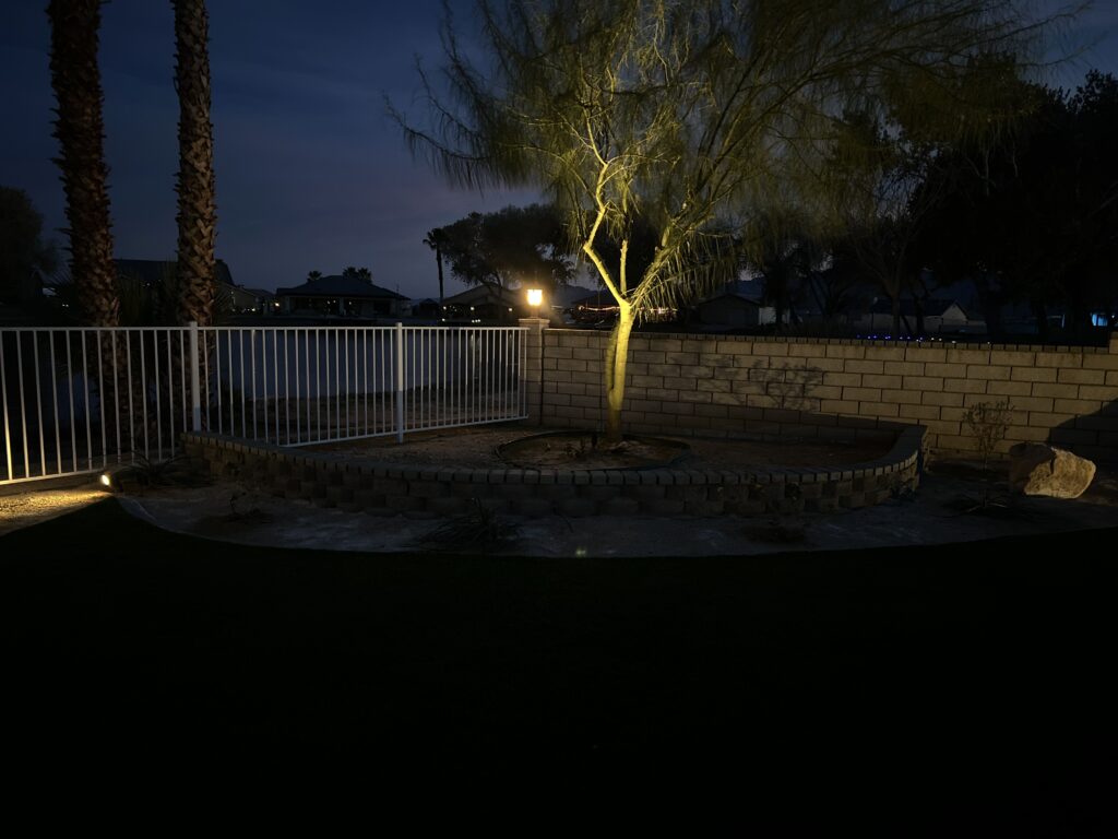 A nighttime view of a landscaped backyard in Silver Lakes featuring a tree illuminated by landscape lighting, a curved retaining wall, white metal fencing, and views of a nearby lake.